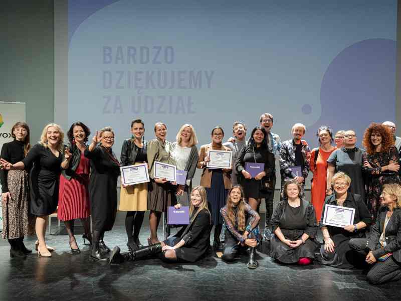 Zdjęcie przedstawia grupę 22 osób stojących na scenie na tle ekranu z napisem "Bardzo dziękujemy za udział". Po prawej stronie stoi baner z logo MagoVox. Część osób stoi, część kuca lub siedzi na ziemi. Uśmiechają się i obejmują, część osób trzyma dyplomy oraz fioletowe albumy z napisem Wzmocnione.