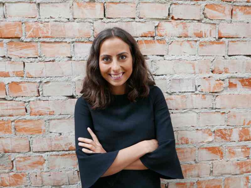 Photo of Pegah Afsharian, Staff for Ashoka Nordics; person with long dark hair and lighter skin smiling at a camera with arms crossed, dressed in a black sweater; background is a wall of aged exposed bricks