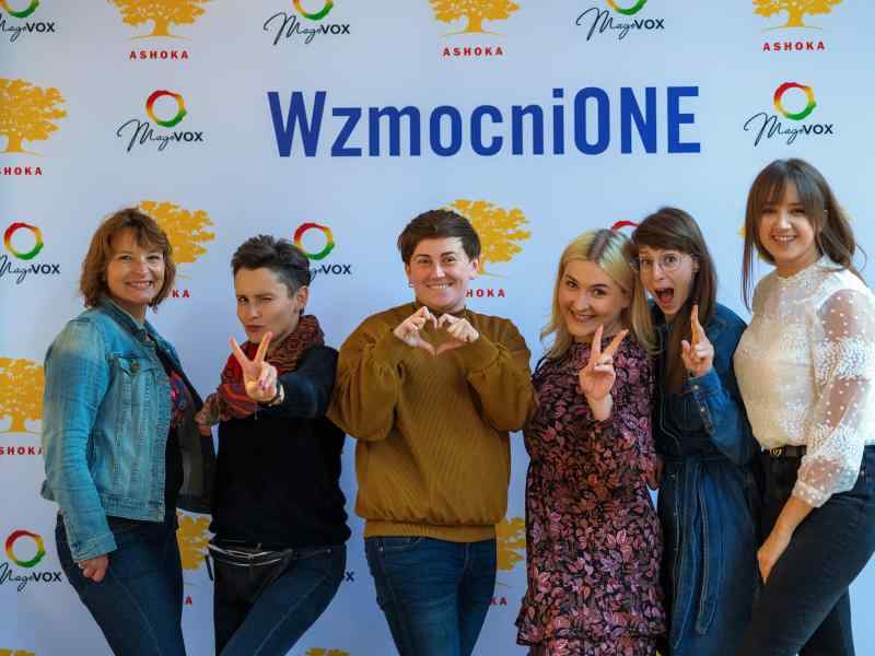 Na tle ścianki wzmocnionej z logo Wzmocnione, Ashoki oraz Magovox pozuje 6 osób w kolorowych ubraniach. Uśmiechają się, 3 pokazują znak peac/wolności palcami, jedna robi serduszko dłońmi.