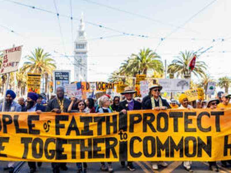 Photo of a faith and climate change protest. Large banner in front saying "People of Faith Rise Together to Protect Our Common Home." Lots of protest signs behind it with a number of people from all different faiths walking on a street. Background of palm trees, and a clock tower