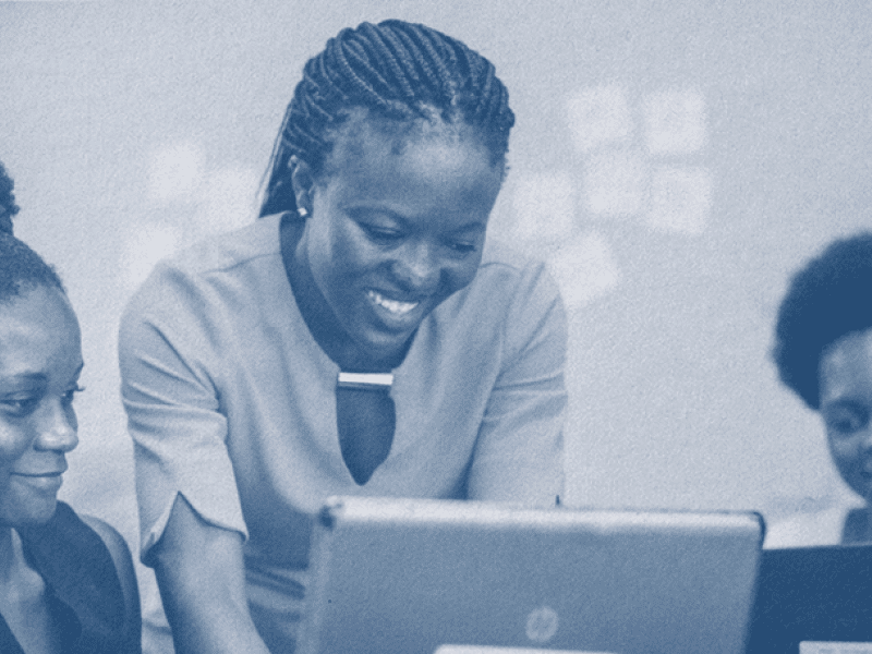 Photo shaded in dark blue. Three people huddle over a few laptops, smiling and looking at the laptops, as if they are working on something together