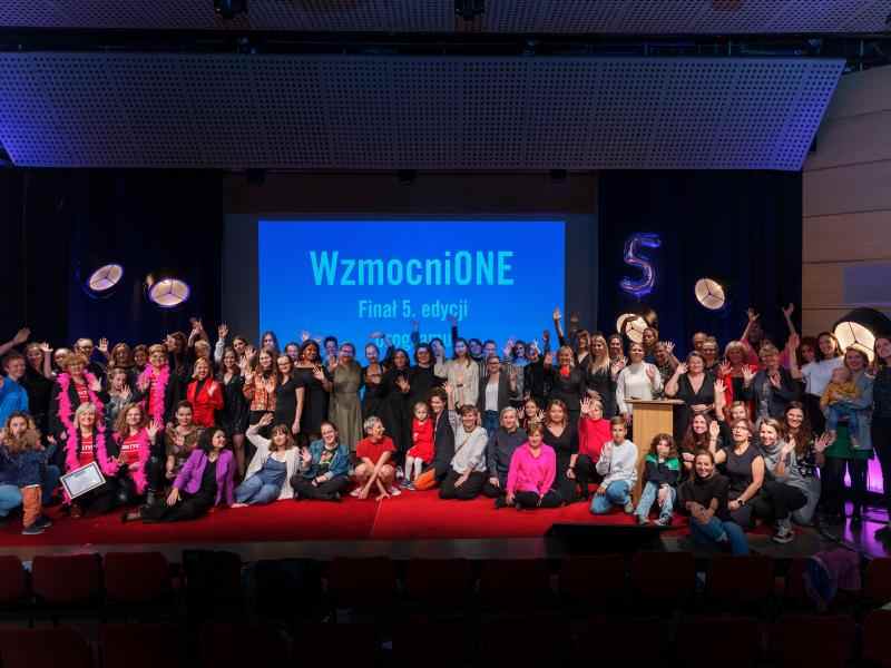 Zdjęcie przedstawia wszystkie osoby obecne na finale 5 edycji programu Wzmocnione na scenie w Muzem Manggha w Krakowie. Osoby uśmiechają się i machają, a za nimi widać rzutnimk z napisem "WzmocniONE. Finał 5 edycji."