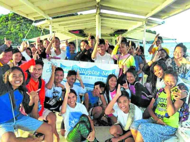More than twenty Filipino youth pose on a beachside pier, holding their hands to their foreheads like sharkfins