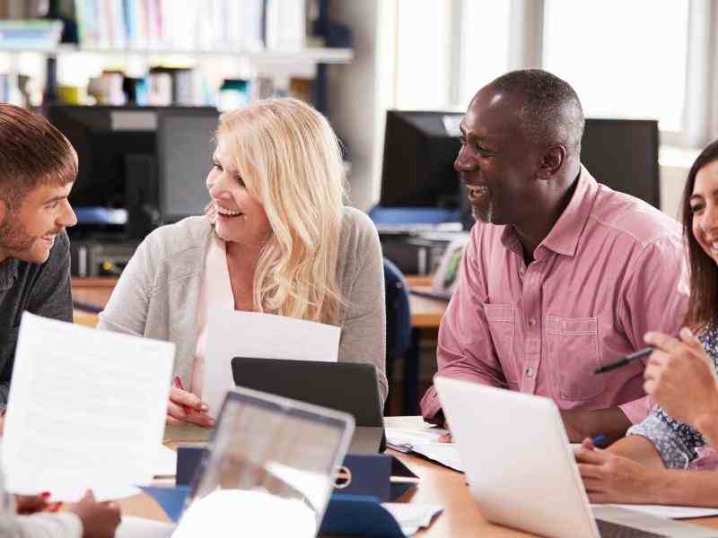 Grupo de pessoas estão conversando e sorrindo em torno de uma mesa em uma biblioteca. Algumas seguram folhas de papel, outras seguram canetas. Há tablets e computadores em cima da mesa