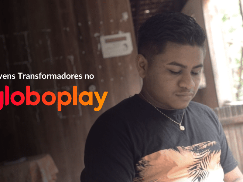 Foto do Jovem Transformador Rian Santos, um jovem quilombola que está olhando para baixo. Ele está em uma sala com paredes de madeira e ao fundo há uma janelaÀ esquerda, há um filtro escuro e lê-se "Jovens Transformadores no Globoplay", sendo a última palavra o logo da plataforma de streaming