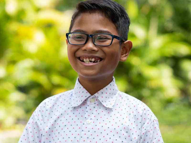 Headshot of AYC Bhre grinning cheerfully wearing a white polkadot shirt on a nature background.
