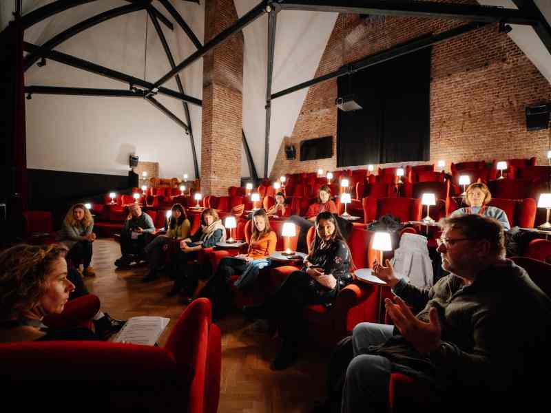 Photo of dark room with red felt chairs, lamps and audience following a session