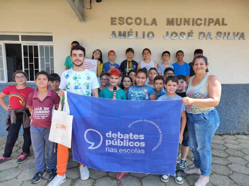 Grupo de crianças reunidas em frente à escola. Dois adultos estão junto com o grupo e seguram a bandeira dos Debates Públicos nas Escolas