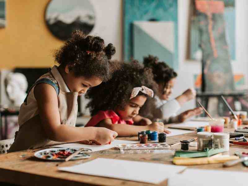 Duas crianças negras estão sentadas a uma mesa em que há tintas. Elas estão desenhando