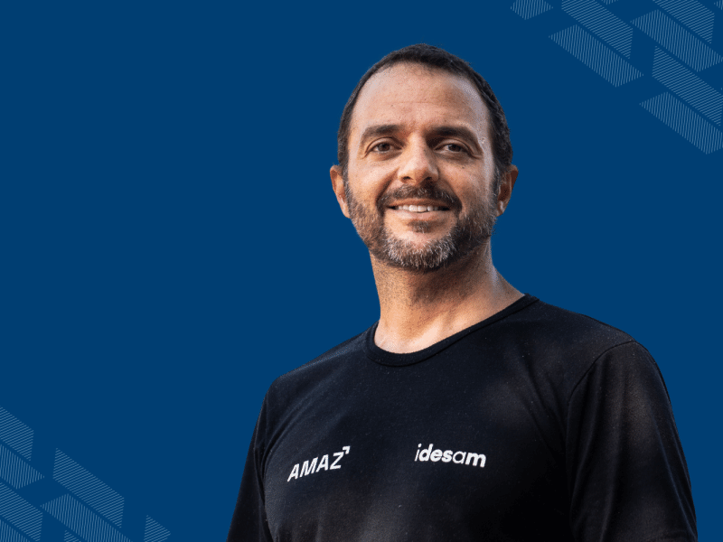 Fundo em azul marinho com grafismos nos cantos. Ao centro, está uma foto de Mariano Cenamo, homem branco com cabelo castanho, curto e barba. Ele veste uma camiseta preta e sorri para a foto