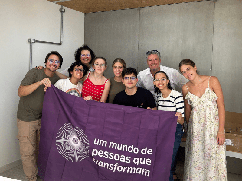 Vê-se uma foto da equipe Ashoka, juntamente com os Jovens Transformadores e Alejandro Villanueva e suas filhas. Na fotografia todos estão segurando a bandeira do movimento Um Mundo de Pessoas que Transformam