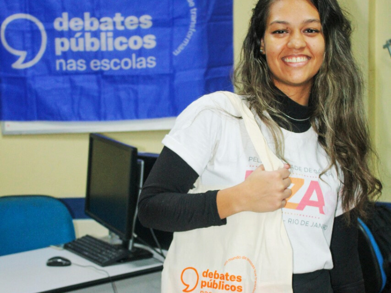 Uma jovem de cabelos longos com mechas loiras sorri para foto segurando a ecobag dos Debates Públicos nas Escolas