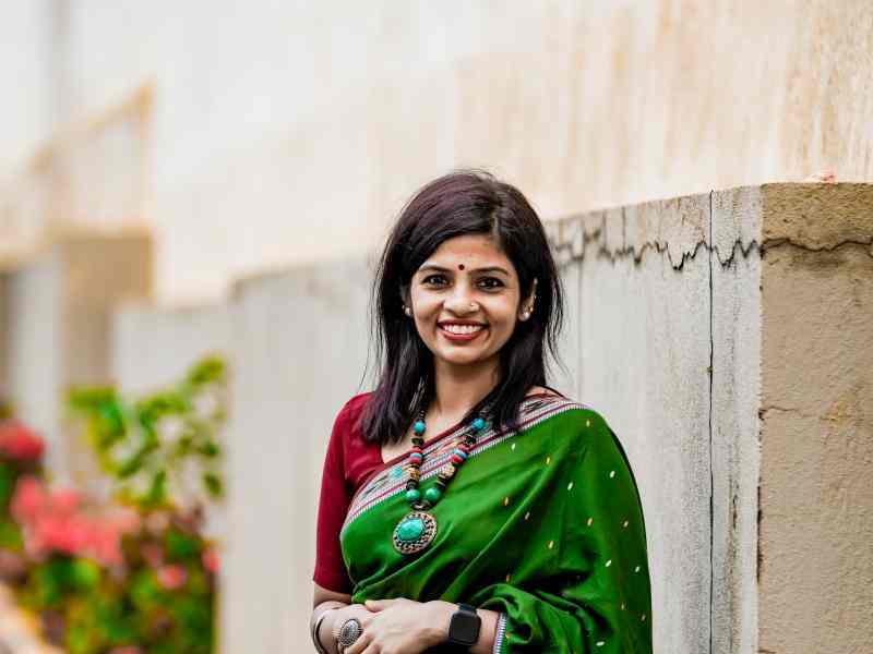 A headshot photo of Shruti Nair posing outside, in front of a stone wall with flowers