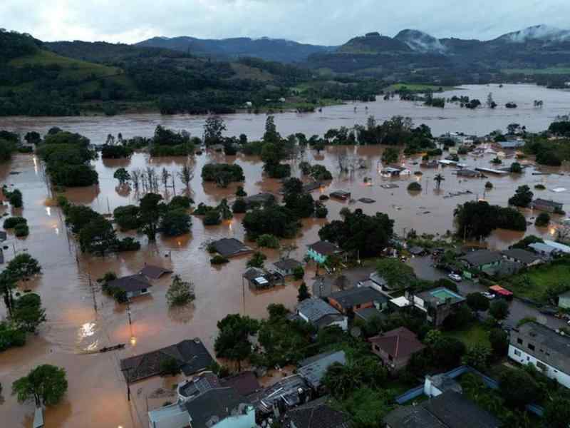 Imagem aérea mostra uma cidade atingidas por enchentes, com a água em tom amarronzado chegando até o telhado das casas e copa das árvores. Ao fundo, vê-se montanhas