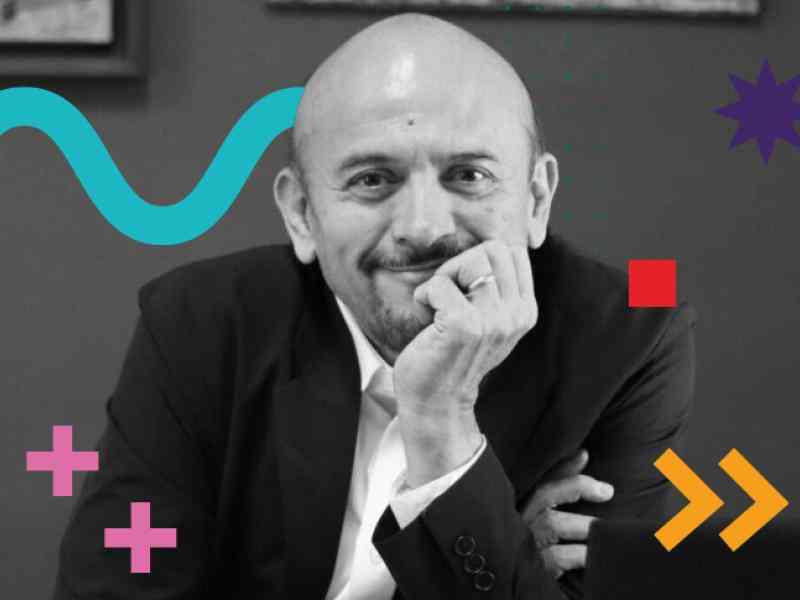 In the photo we can see Mauro - a Latin man, bald, wearing a suit and dress shirt. The photo is in black and white, and we have colorful graphic features added to the photo: a blue zigzag curve, a red square, a purple 8-pointed star, green dots, two pink plus signs and two orange arrows. 