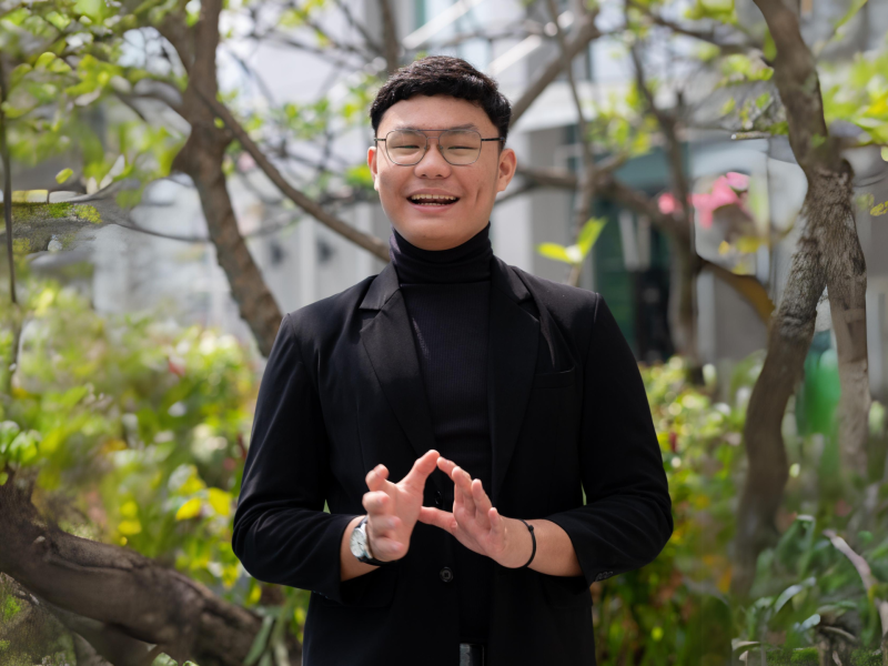 Headshot of AYC Atha smiling at the camera wearing glasses and a black suit on a nature background.