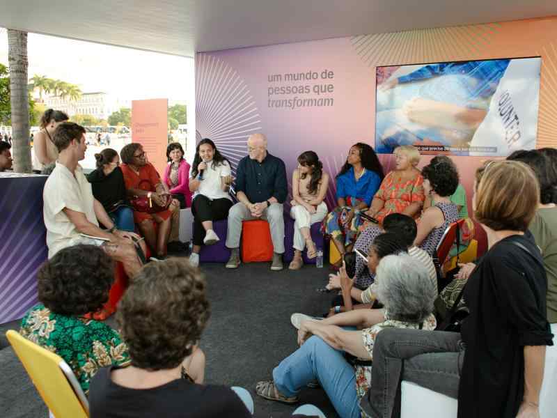 Grupo de pessoas reunida em um estande ao ar livre. Elas estão sentadas em roda, em pufes, bancos e cadeiras de praia. Ao fundo, há um painel colorido onde se lê "um mundo de pessoas que transformam" com uma televisão ao lado