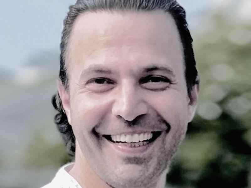 Profile photo of Ashoka Framework Change Staff Edward Edilbi. Photo of person with long slick backed hair smiling at the camera; background is of blurred out trees