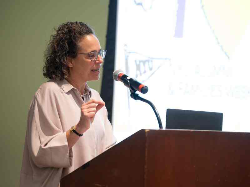 Photo of Diana Wells accepting GSAS Distinguished Alumni Achievement Award. Person with dark curly shoulder length hair and glasses speaking into a microphone attached to a dark brown podium. Dressed in light pink blouse; background is the front of a what appears to be a classroom or auditorium