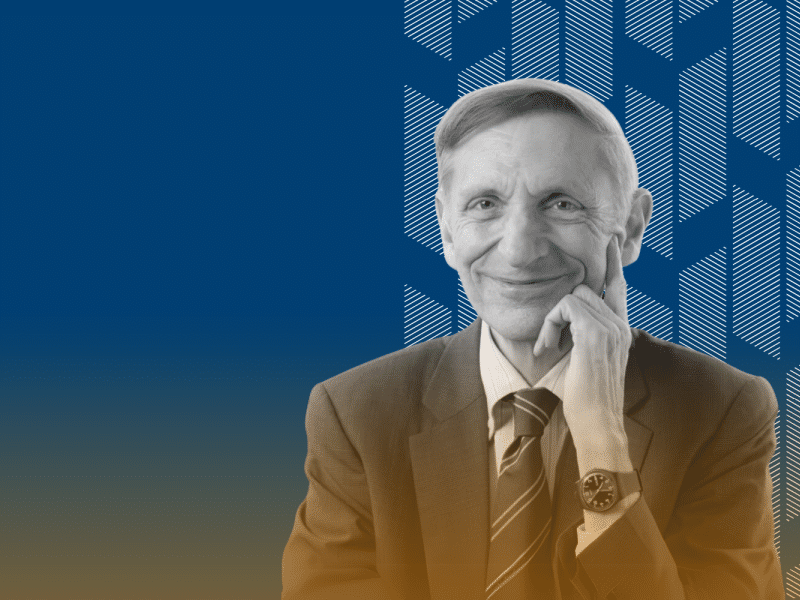 Fundo azul marinho com grafismos brancos à direita. Sobre o padrão de grafismos, há uma foto em preto e branco de Bill Drayton, homem branco de cabelos claros e curtos. Ele sorri levemente e veste um terno com gravata listrada e um relógio no pulso esquerdo. Na parte inferior, há um gradiente laranja
