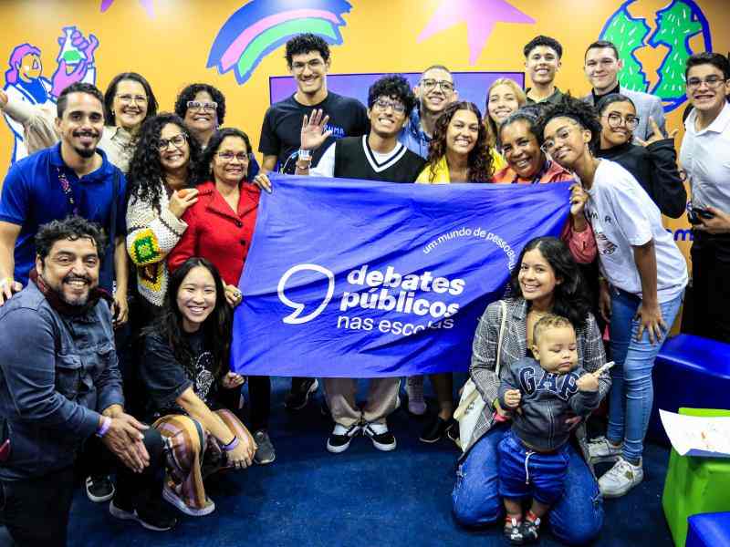 Grupo de pessoas de diferentes idades reunidas em um stand ao ar livre. Em destaque, elas seguram a bandeira dos Debates Públicos nas Escolas. Ao fundo, há um painel laranja com ilustrações coloridas.