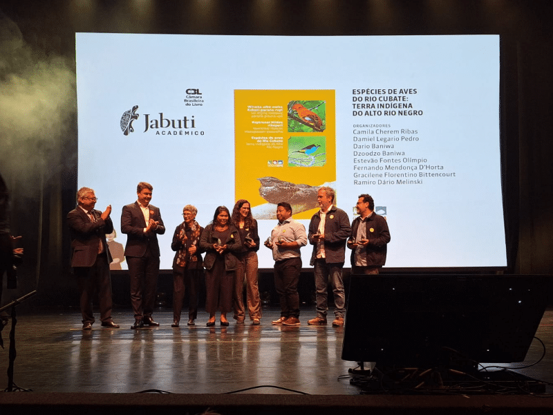 Pessoas em pé sobre um palco, algumas aplaudindo, outras sorrindo e segurando troféus. Ao fundo, um telão projeta a capa do livro “Espécies de Aves do Rio Cubaté: Terra Indígena do Alto Rio Negro”, com ilustrações de aves coloridas. No canto esquerdo da projeção, aparecem os logos da Câmara Brasileira do Livro e do Prêmio Jabuti Acadêmico.