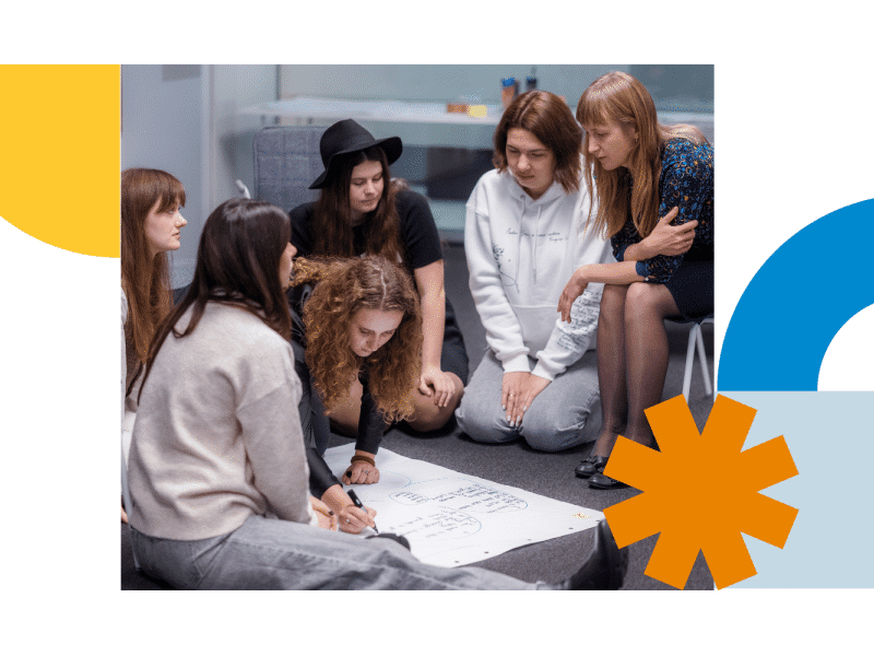 A group of six people is sitting on the floor in a modern indoor setting, gathered closely around a large sheet of white paper placed in the center. The paper appears to have diagrams or handwritten notes on it. The individuals are dressed casually; one person is wearing a black hat, and another is in a white sweatshirt. They are leaning forward, actively engaged with the content on the paper, suggesting a collaborative activity or brainstorming session.