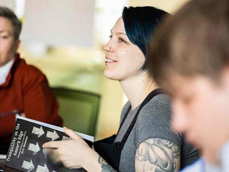Three people sitting in a classroom setting; one person with lighter skin and dark blue hair smiling to the left of the photo.