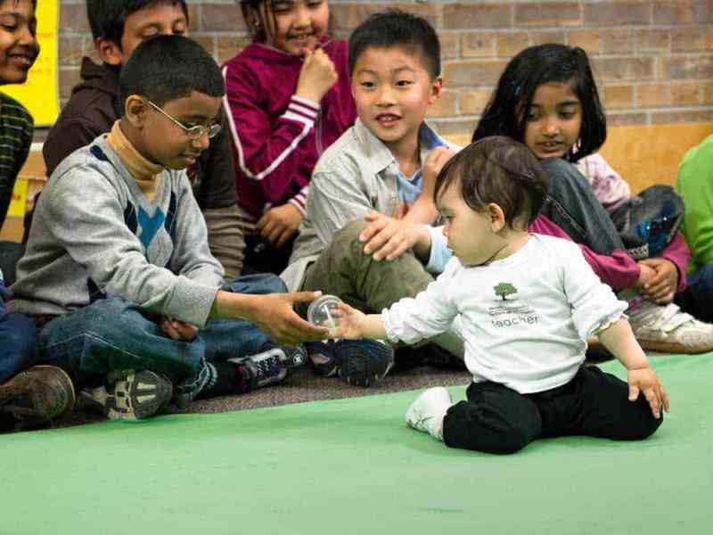 Baby handing an elementary school aged child an object while other students look on; located in a classroom