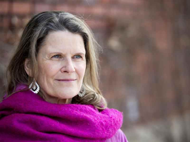 Photo of Canada Fellow Rachel Kiddell-Monroe. Photo of a person with longer brown hair and lighter skin smiling to the right of the camera. Dressed in a pink scarf; background of a brick wall.