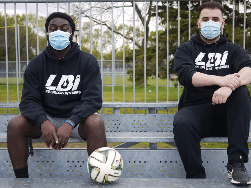 Two soccer/football players seated in the bleachers wearing "Les Ballons Intensifs" hoodies. Both are masked.