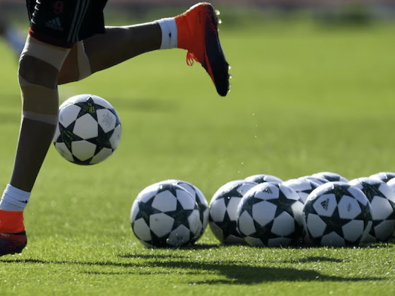 Photo of bottom half of soccer player's body  running on a field; next to him are a number of soccer balls