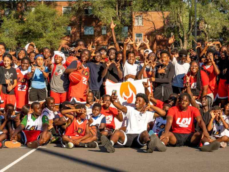 Banner of youth participants involved with Les Ballons Intensifs; about 100 youth of diverse backgrounds lined up in three rows on a basketball court smiling at the camera