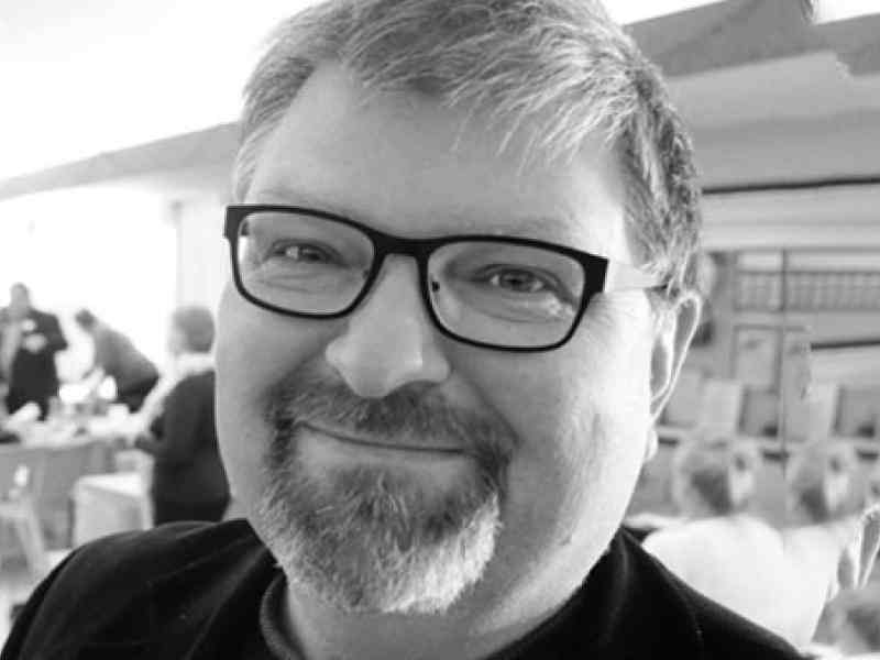 Black and white Headshot of Canada Fellow Paul Born. Person with glasses, short light hair and a goatee smiling at the camera. Background a conference room