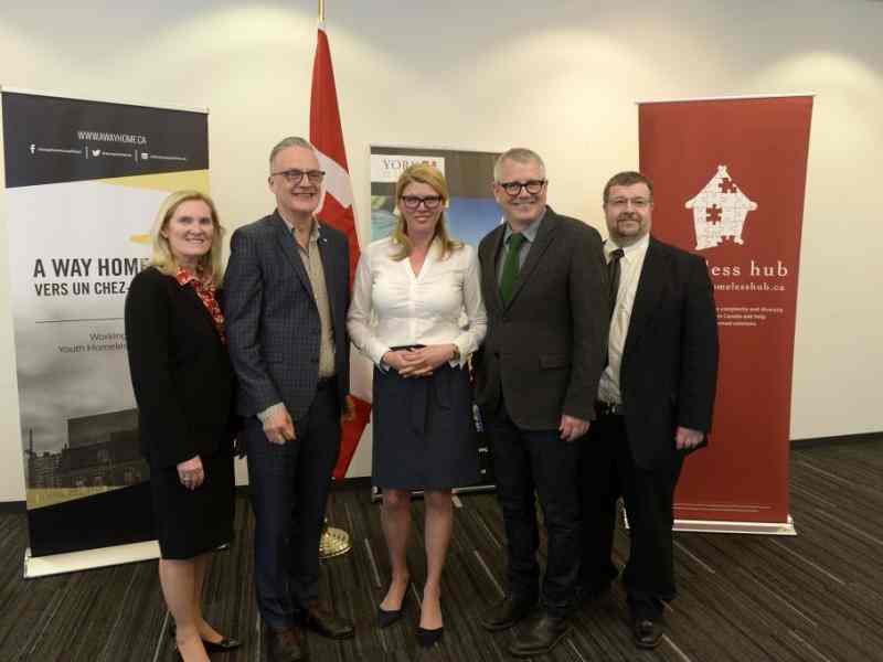 Five people dressed in suits smiling at the camera; backdrop of a few posters relating to the Federal funding for youth homelessness in Canada