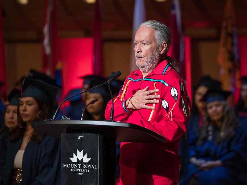 Person with grey hair and beard dressed in a red formal long shirt with indigenous art on it holding hand over heart and speaking to a graduation on a stage