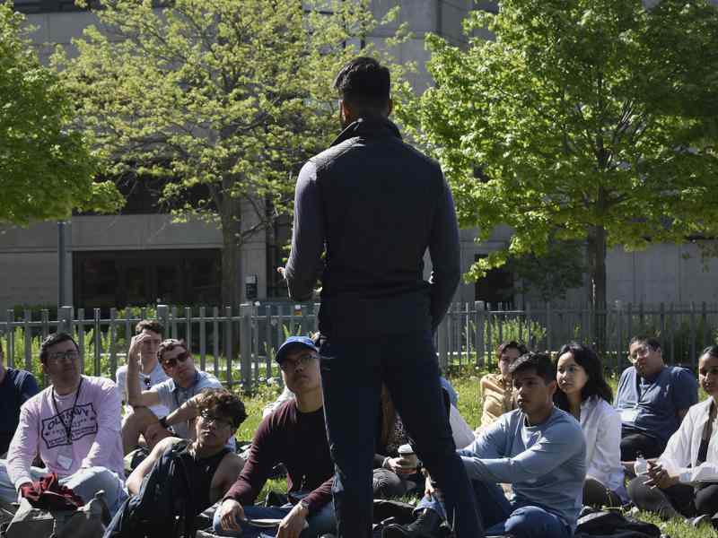 Person standing up and speaking in front of a seated group of people in a field