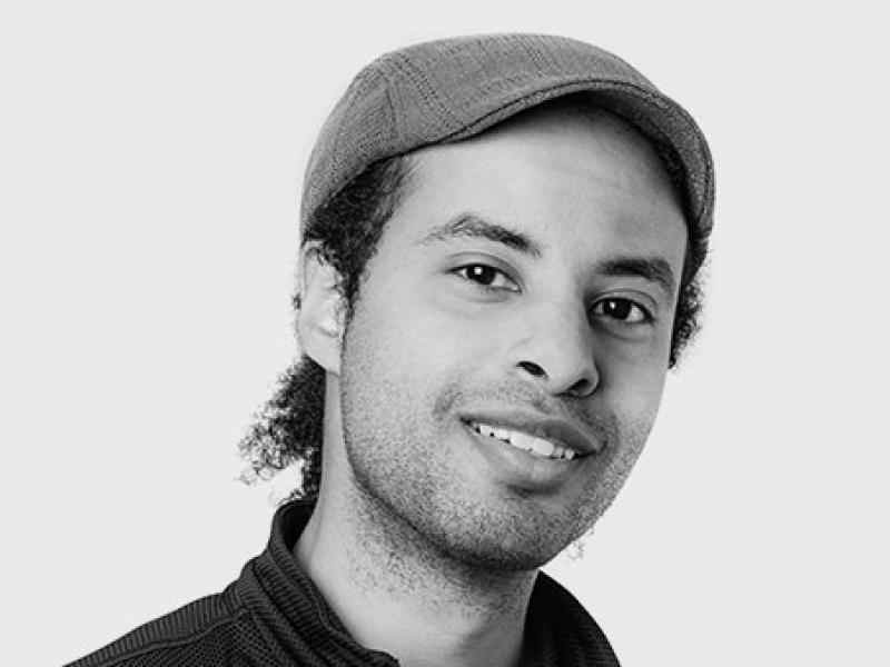 Person with fedora and short dark hair smiling at the camera. Photo in black and white