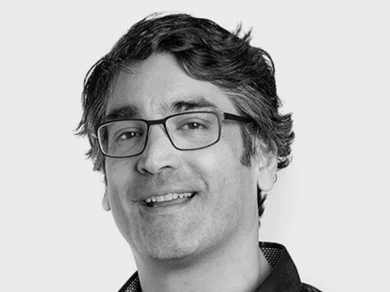Headshot of Anil Patel, Ashoka Fellow in Canada. Black and white photo of a person with wavy hair and glasses smiling at the camera. Background is white wall