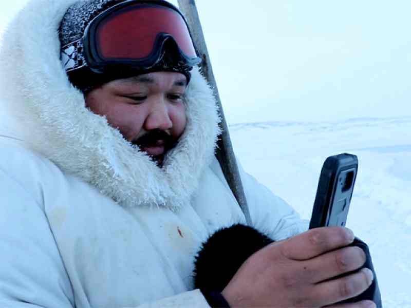 Person with white parka and ski goggles on using an app on a phone; background is a white snowy tundra