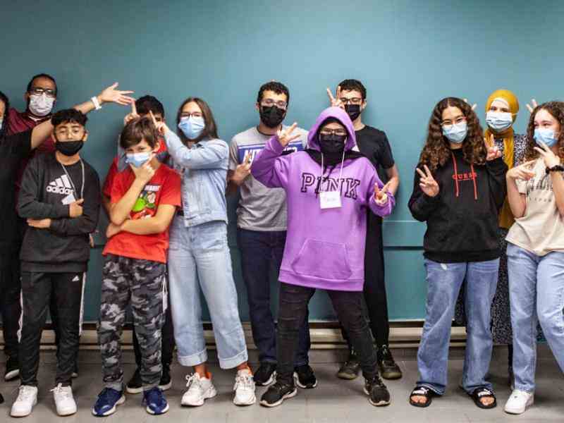 Group of kids with masks on smiling at the camera in a hallway; wallpaper is bright green-blue color