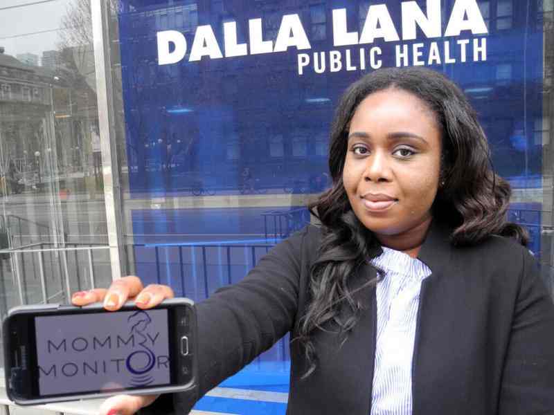 Person with short dark hair and brown skin smiling at the camera, dressed in a business suit. Showing a cell phone horizontally to the camera that has a logo for "Mommy Monitor" on it. Background is Dalla Lana Public Health school at U Toronto
