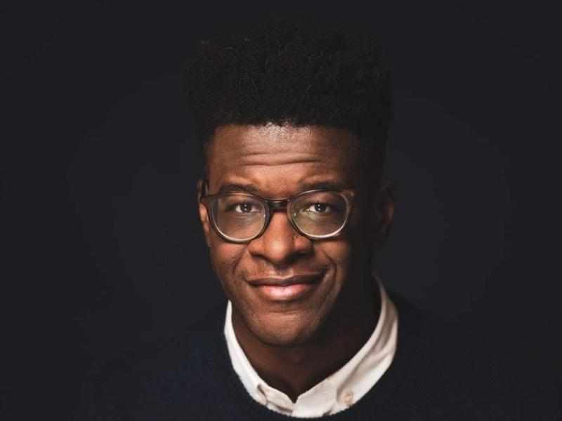 Person with short dark hair and dark brown skin with glasses smiling at the camera, dressed in a black sweater and white collared shirt