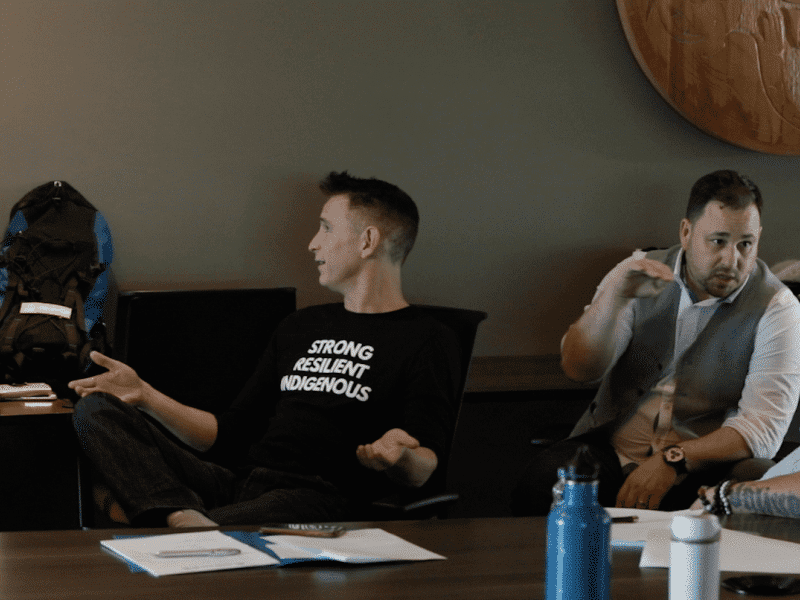 Four people discussing with each other in 1:1 pairs on two couches that are perpendicular to each other, in an indoor room with dark green wallpaper