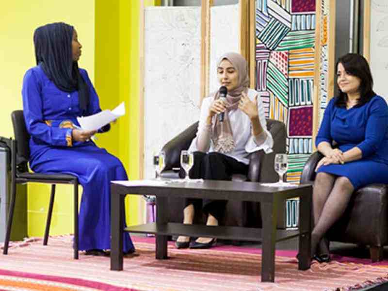 Two young Muslim women speaking on a stage as part of a panel. Background a lifted stage and a few banners