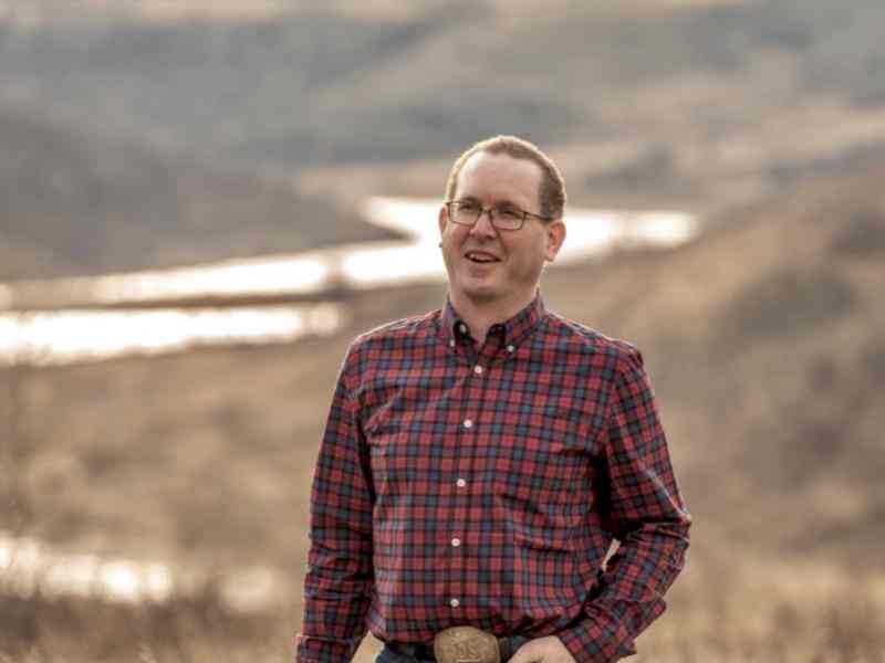 Photo of Sean Mcgrath, rancher in Canada with ALUS. Person with short dark hair, glasses, and a flannel shirt smiling at the camera; background is a river between hills