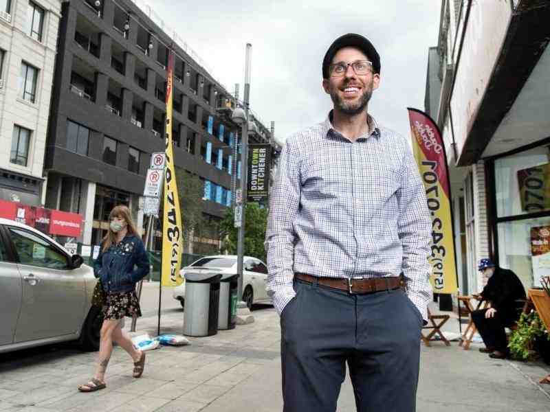 Person with a news cap on dressed in formal dresswear smiling at the camera backdrop of a sidewalk leading down a street behind the person