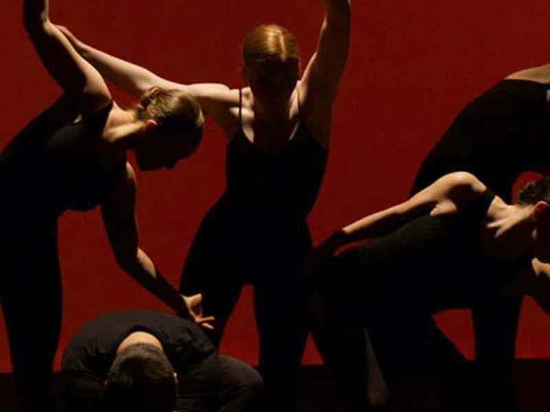 Picture of 5 dancers dressed in black tight dancing attire in a low light with red background arranged closely to each other mid choregraphy