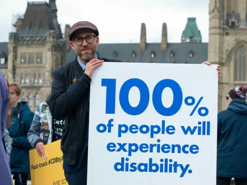 Person with a sign that says 100% of people will experience disability, background an old building below a skyline