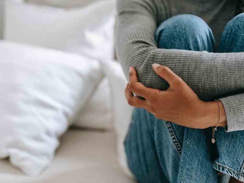 Person with femme attributes holding her knees on a bed; face off camera